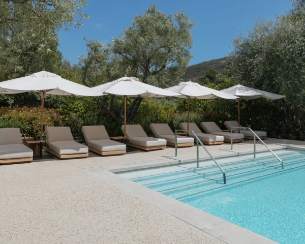 Row of beige pool loungers under white umbrellas by a blue pool with trees and hills in the background.