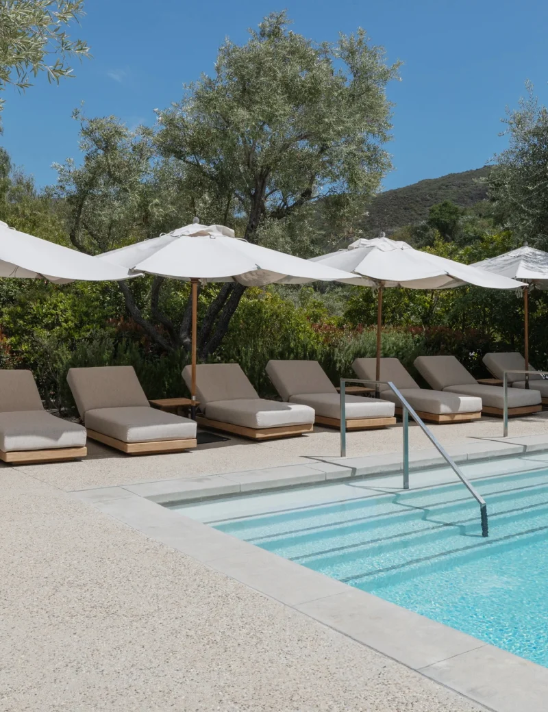 Row of beige pool loungers under white umbrellas by a blue pool with trees and hills in the background.