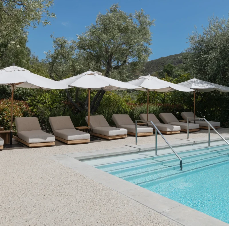 Row of beige pool loungers under white umbrellas by a blue pool with trees and hills in the background.