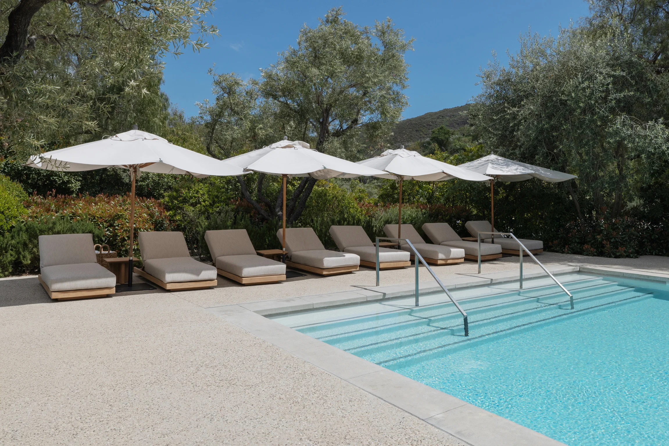 Row of beige pool loungers under white umbrellas by a blue pool with trees and hills in the background.