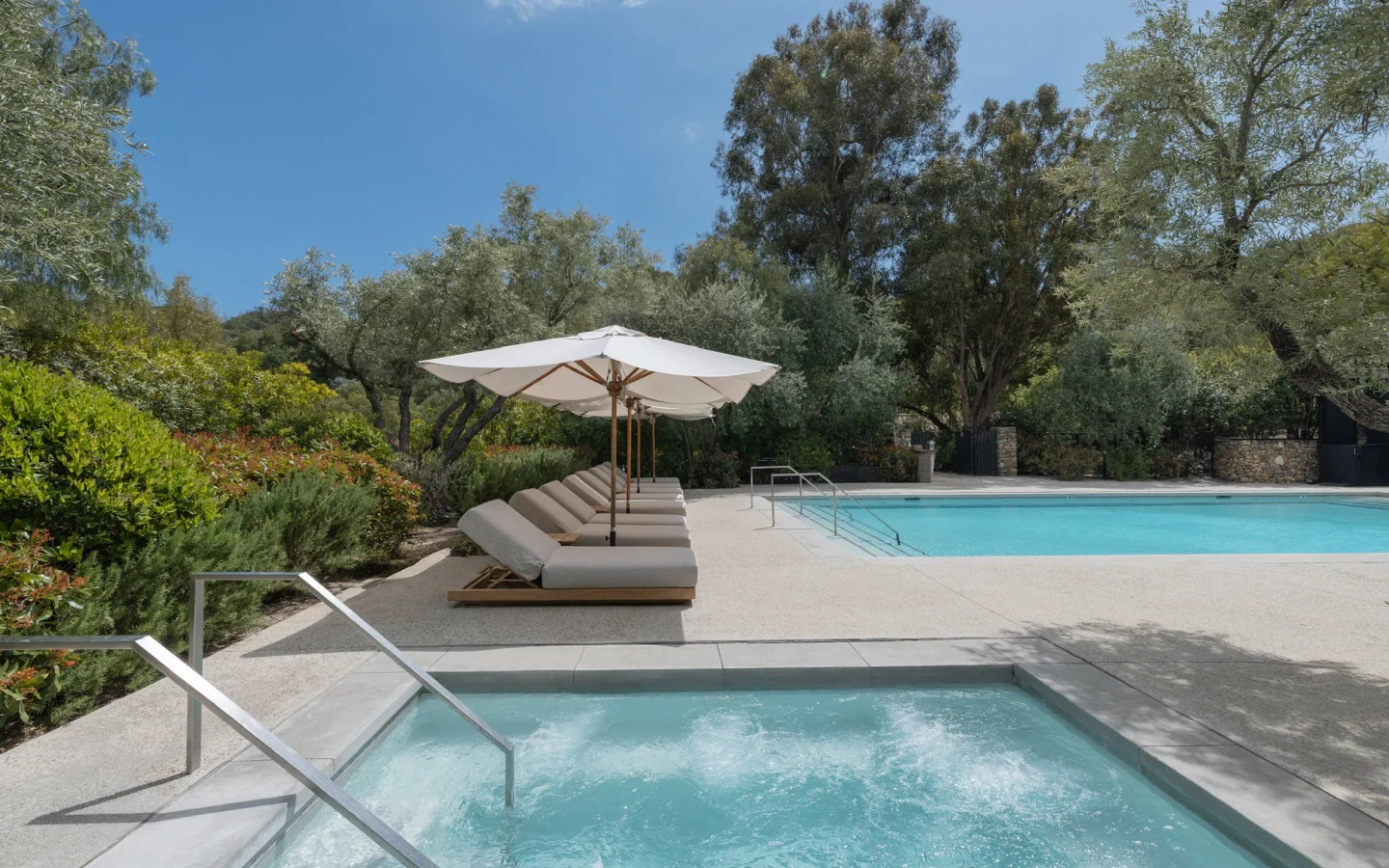 Poolside with white umbrellas and cushioned lounge chairs beside a clear blue pool amid lush greenery and trees.