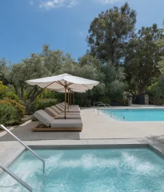 Poolside with white umbrellas and cushioned lounge chairs beside a clear blue pool amid lush greenery and trees.