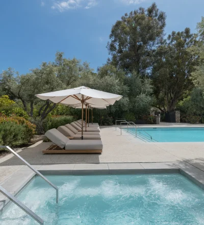 Poolside with white umbrellas and cushioned lounge chairs beside a clear blue pool amid lush greenery and trees.