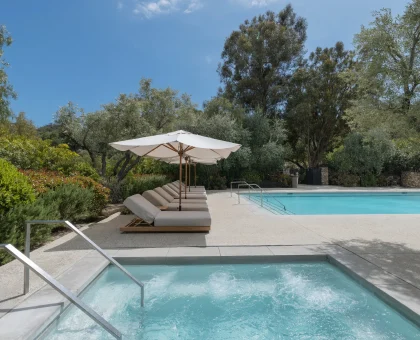 Poolside with white umbrellas and cushioned lounge chairs beside a clear blue pool amid lush greenery and trees.