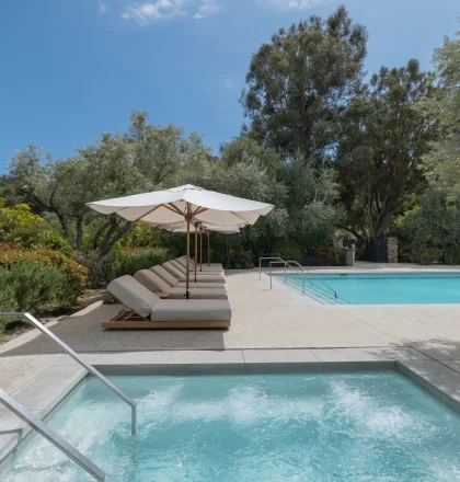 Poolside with white umbrellas and cushioned lounge chairs beside a clear blue pool amid lush greenery and trees.