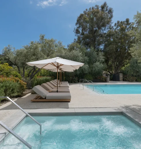 Poolside with white umbrellas and cushioned lounge chairs beside a clear blue pool amid lush greenery and trees.