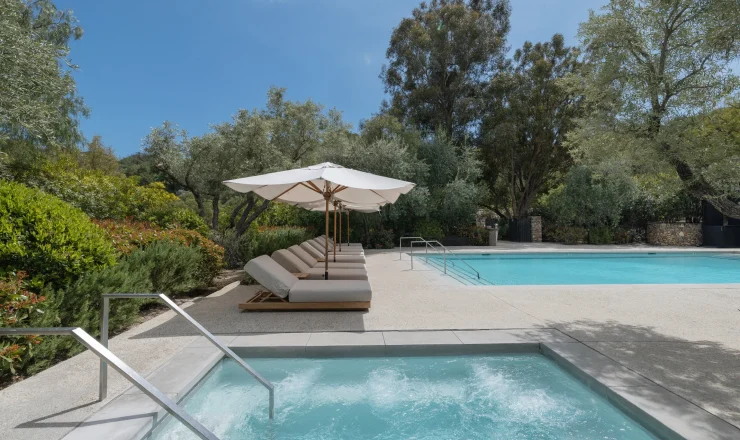 Poolside with white umbrellas and cushioned lounge chairs beside a clear blue pool amid lush greenery and trees.