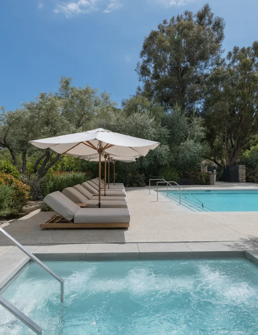 Poolside with white umbrellas and cushioned lounge chairs beside a clear blue pool amid lush greenery and trees.