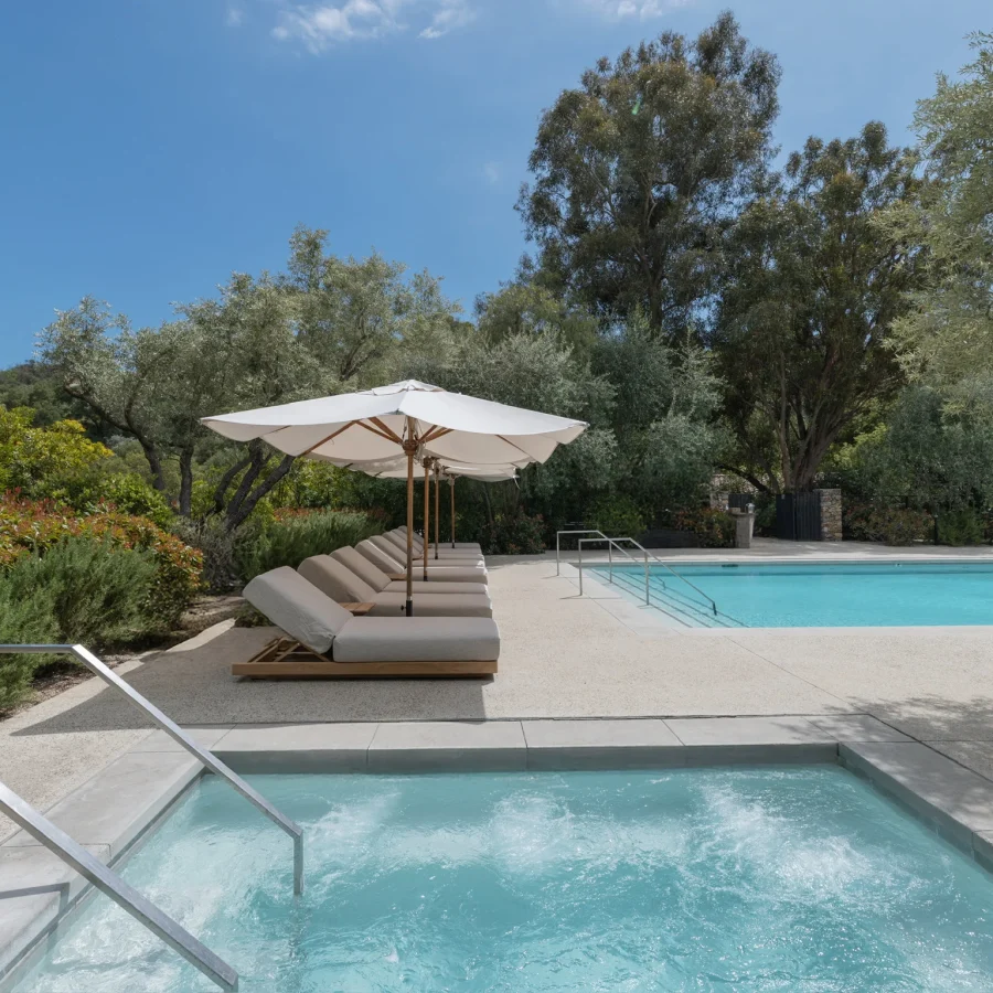 Poolside with white umbrellas and cushioned lounge chairs beside a clear blue pool amid lush greenery and trees.