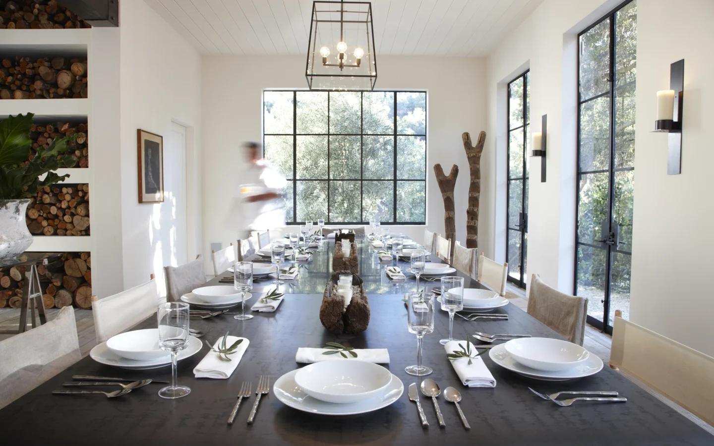 Long dining table set for a meal in a bright, modern room with large grid windows and stacked firewood shelves on the left.