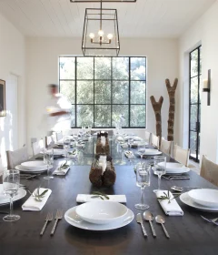 Long dining table set for a meal in a bright, modern room with large grid windows and stacked firewood shelves on the left.