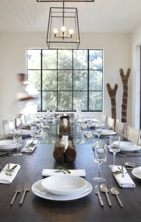 Long dining table set for a meal in a bright, modern room with large grid windows and stacked firewood shelves on the left.