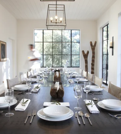 Long dining table set for a meal in a bright, modern room with large grid windows and stacked firewood shelves on the left.