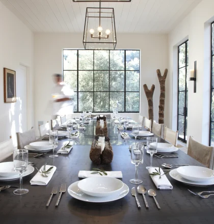 Long dining table set for a meal in a bright, modern room with large grid windows and stacked firewood shelves on the left.