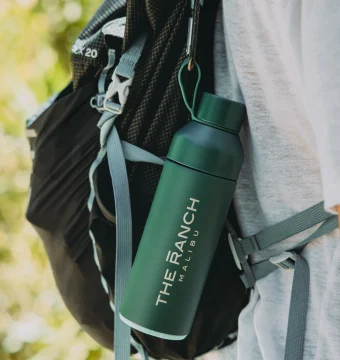 Close-up of a dark hiking backpack with a green water bottle in a side pocket and metal clips hanging from a strap, outdoors with a blurred green background.