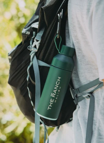 Close-up of a dark hiking backpack with a green water bottle in a side pocket and metal clips hanging from a strap, outdoors with a blurred green background.