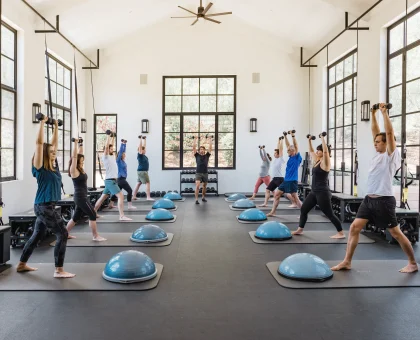Group fitness class in a bright studio with participants lifting dumbbells overhead while standing on mats, surrounded by balance domes and large windows.
