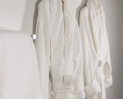 Two white bathrobes hanging on metal hooks on a light wall in a bathroom.