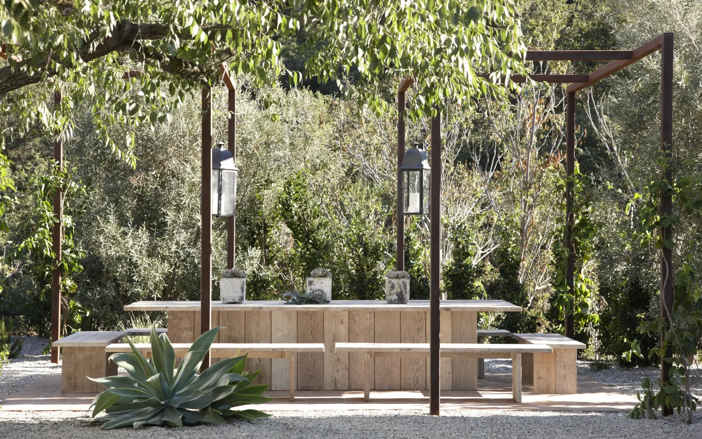 Wooden outdoor dining table and benches under a pergola, with lanterns and lush greenery around them.