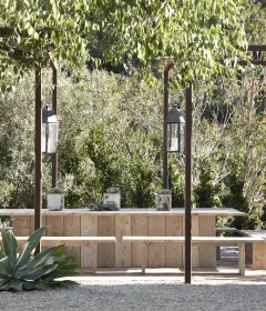 Wooden outdoor dining table and benches under a pergola, with lanterns and lush greenery around them.
