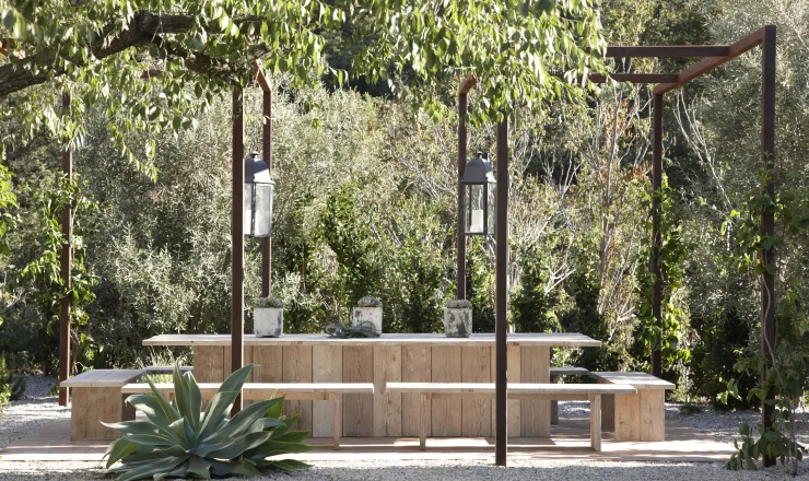 Wooden outdoor dining table and benches under a pergola, with lanterns and lush greenery around them.