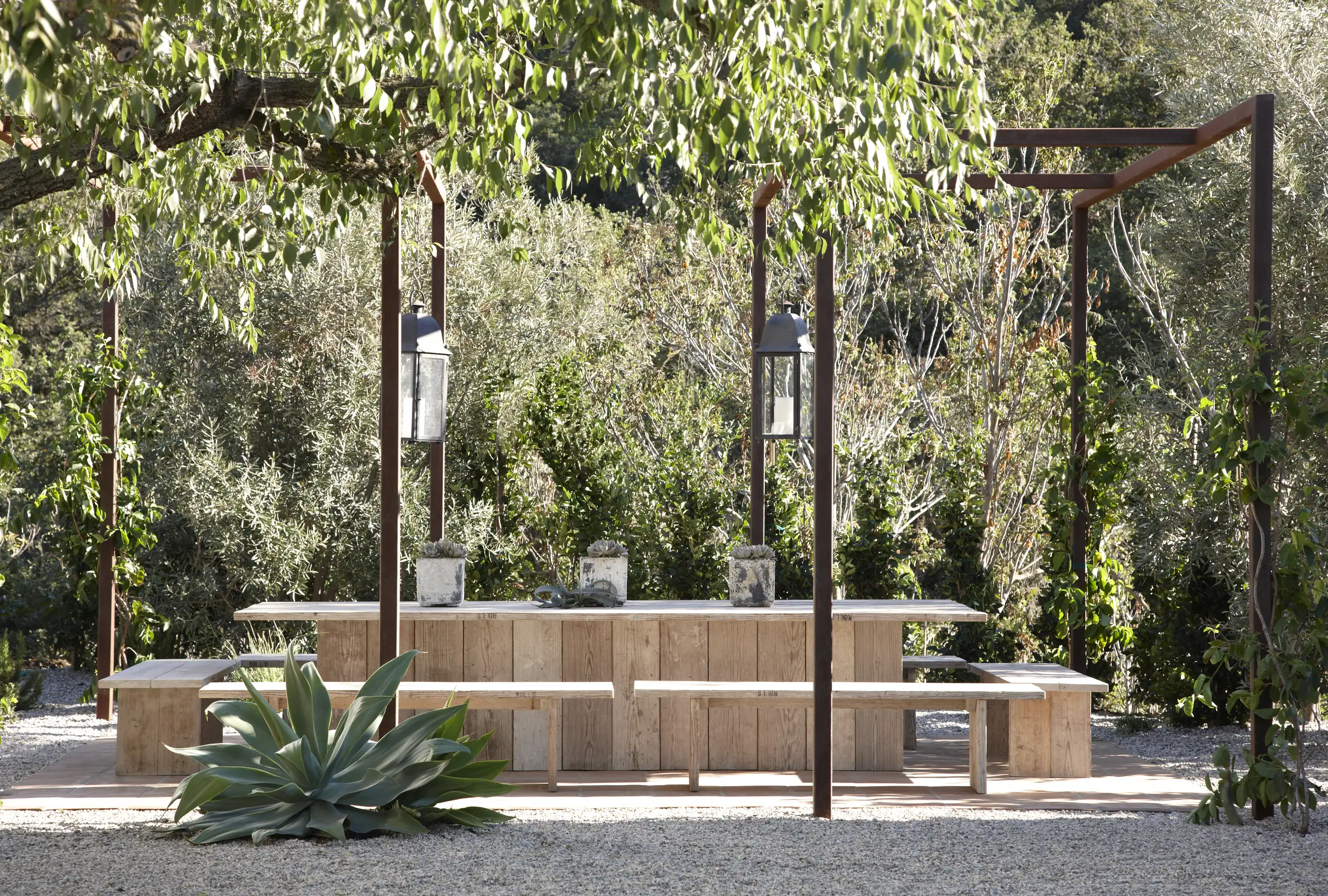 Wooden outdoor dining table and benches under a pergola, with lanterns and lush greenery around them.