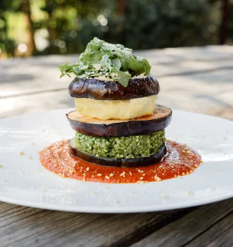 Layered grilled eggplant stack with pesto, greens, and grated cheese on a white plate with red sauce.