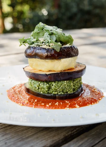 Layered grilled eggplant stack with pesto, greens, and grated cheese on a white plate with red sauce.