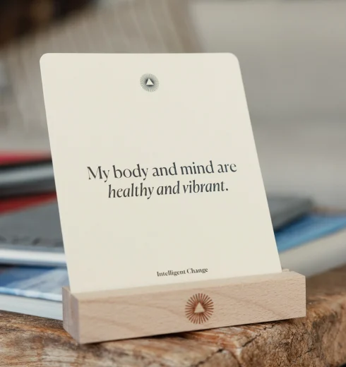 Card on a wooden stand displaying the phrase: 'My body and mind are healthy and vibrant,' with a small circular sunburst logo above and 'Intelligent Change' at the bottom edge.
