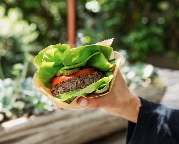 Person holding a burger wrapped in paper with lettuce and tomato, outdoors with greenery in the background.