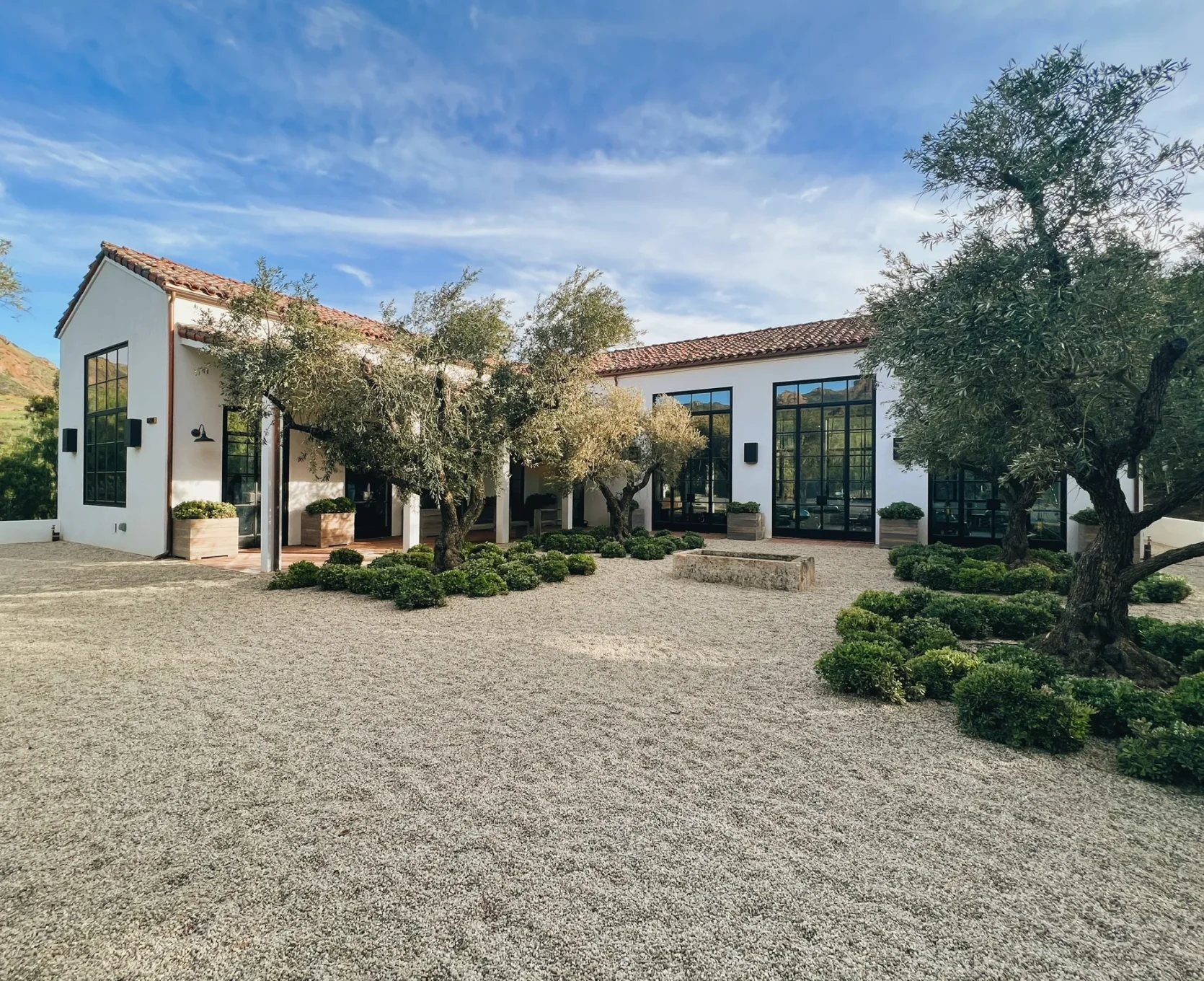White modern building with large black-framed windows, red-tile roof, and olive trees in a gravel courtyard. The scene is sunny and spacious.