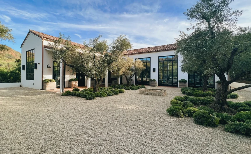 White modern building with large black-framed windows, red-tile roof, and olive trees in a gravel courtyard. The scene is sunny and spacious.
