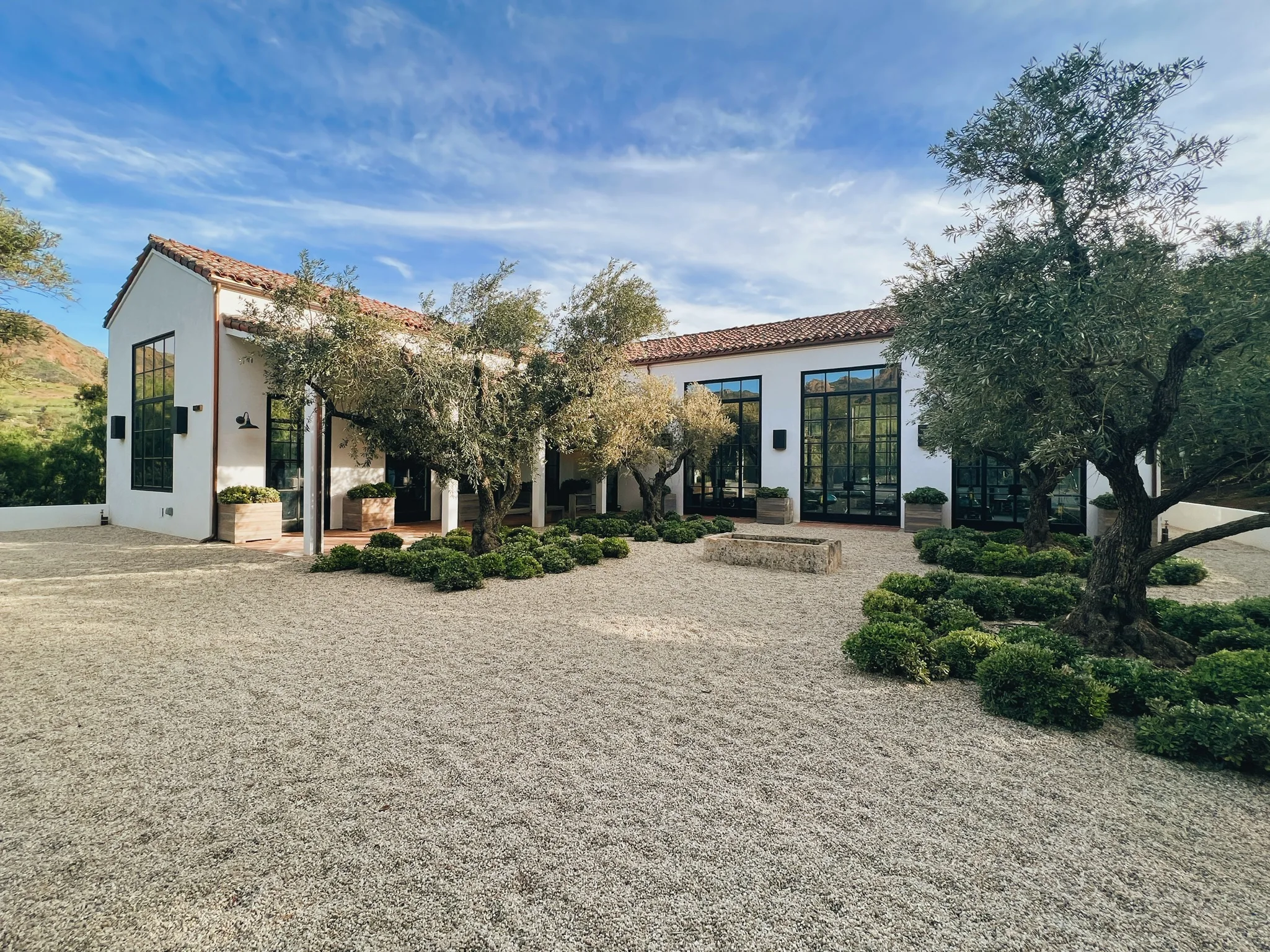 White modern building with large black-framed windows, red-tile roof, and olive trees in a gravel courtyard. The scene is sunny and spacious.