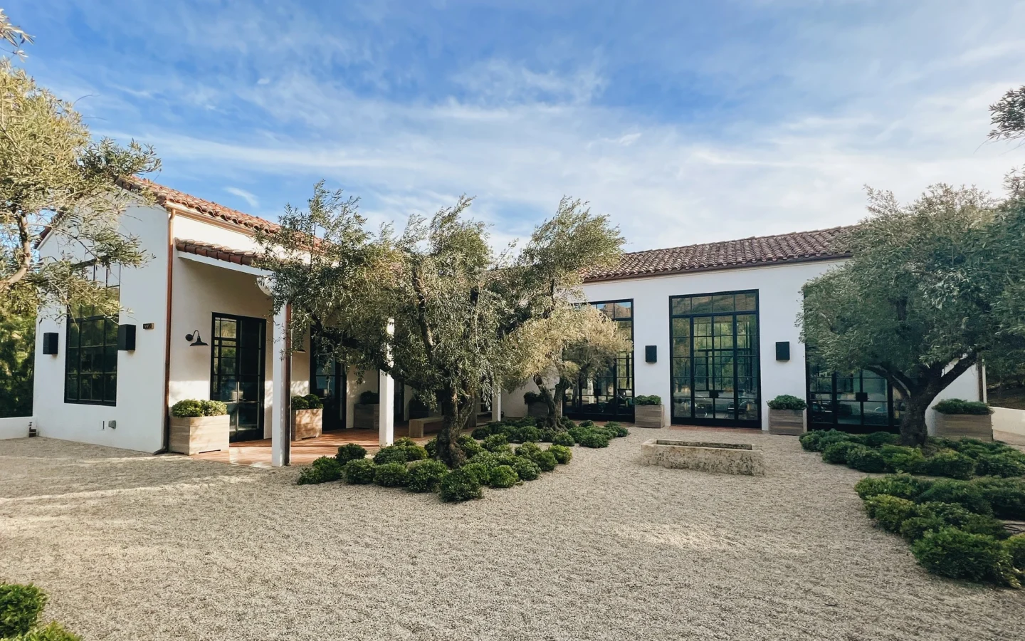 White modern building with large black-framed windows in a gravel courtyard, olive trees, and a blue sky above.