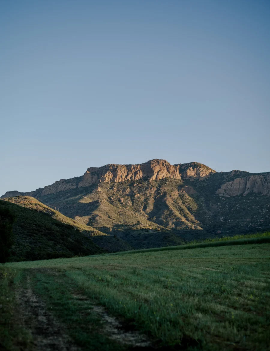 Sunlit rocky hills rise from a green meadow under a clear blue sky.