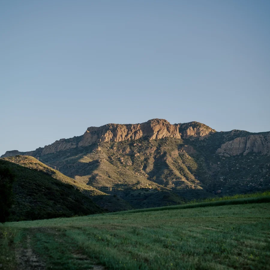 Sunlit rocky hills rise from a green meadow under a clear blue sky.