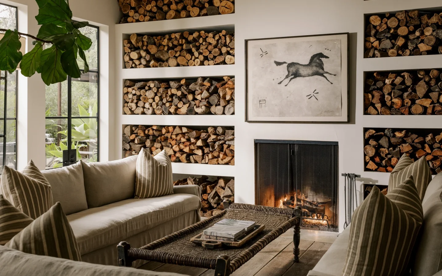 Cozy living room with built-in log storage, beige sofas, and a lit fireplace beneath a black geometric chandelier.