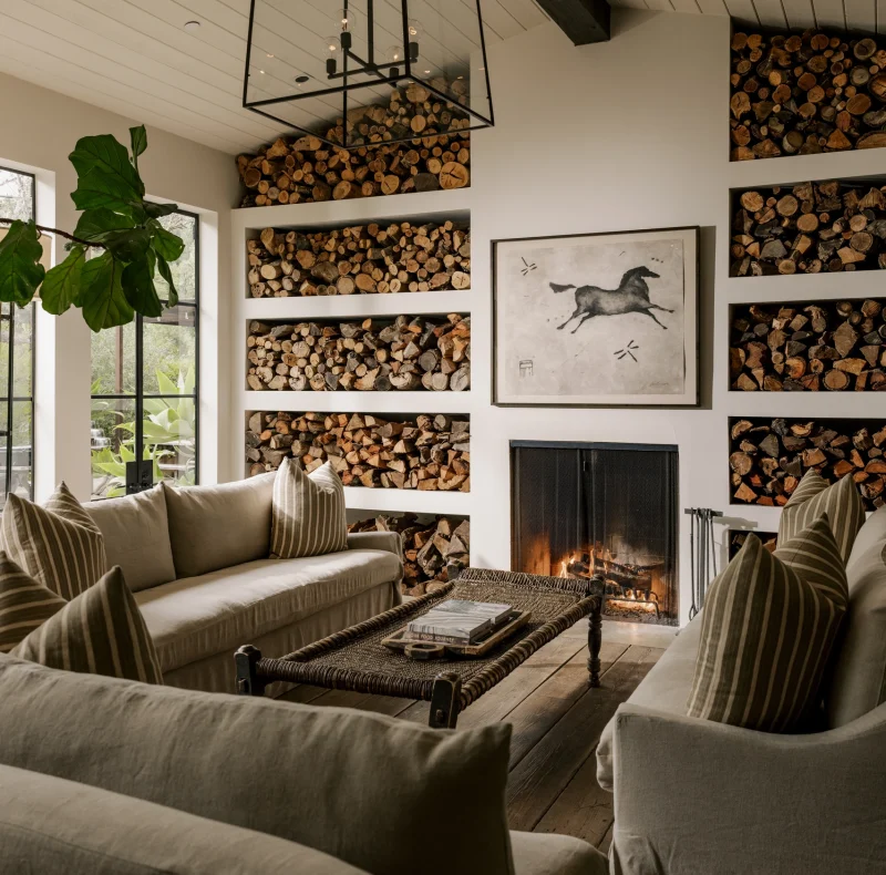 Cozy living room with built-in log storage, beige sofas, and a lit fireplace beneath a black geometric chandelier.