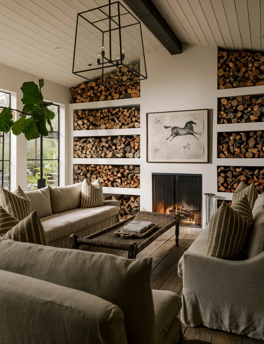 Cozy living room with built-in log storage, beige sofas, and a lit fireplace beneath a black geometric chandelier.