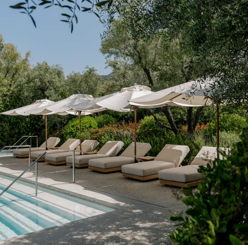Row of beige lounge chairs beneath white umbrellas beside a sunlit outdoor pool.