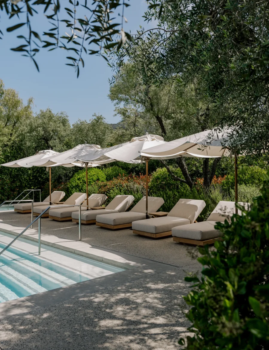Row of beige lounge chairs beneath white umbrellas beside a sunlit outdoor pool.