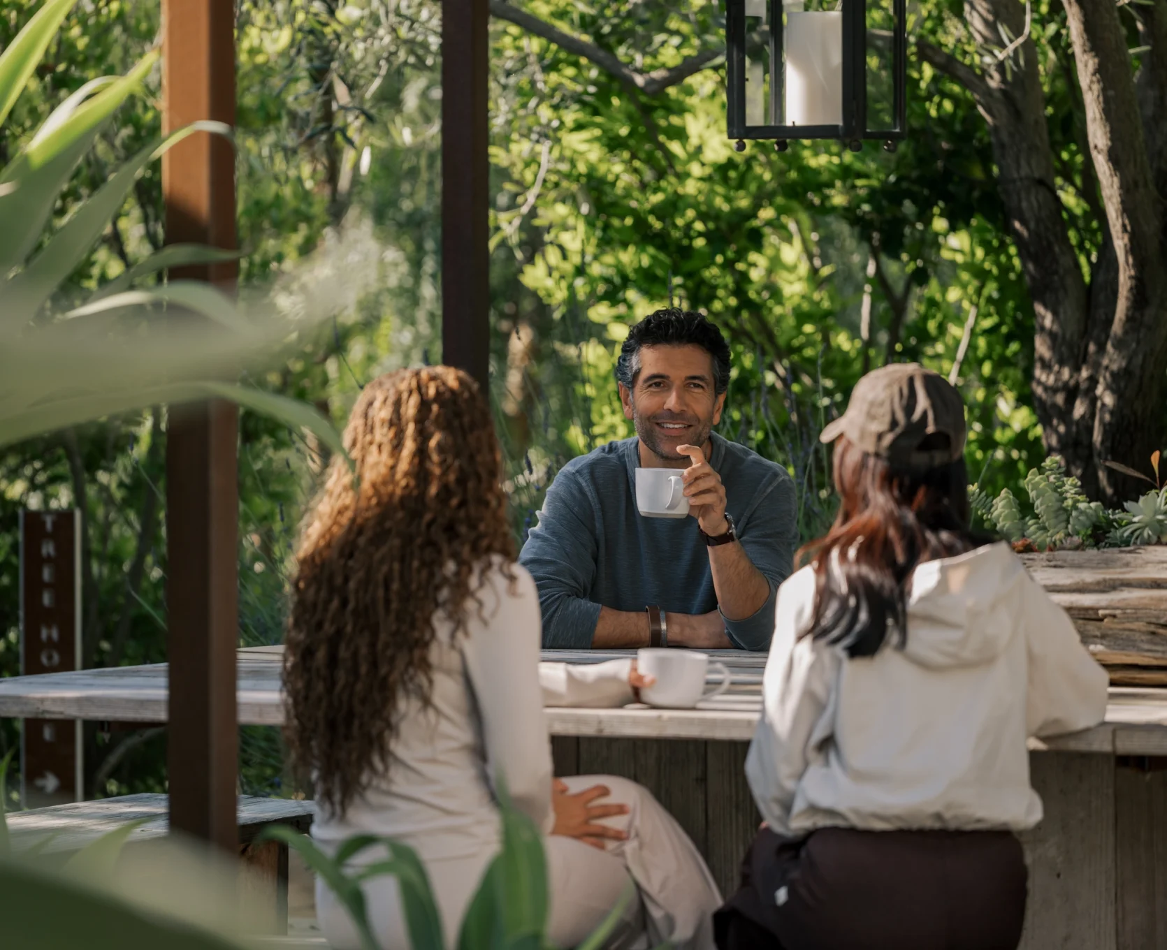 Group of people seated outdoors around a wooden table, chatting and sipping coffee in a sunny garden setting.