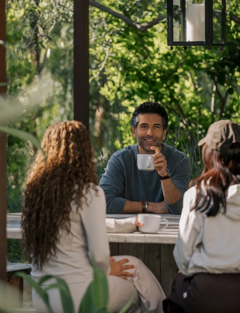 Group of people seated outdoors around a wooden table, chatting and sipping coffee in a sunny garden setting.