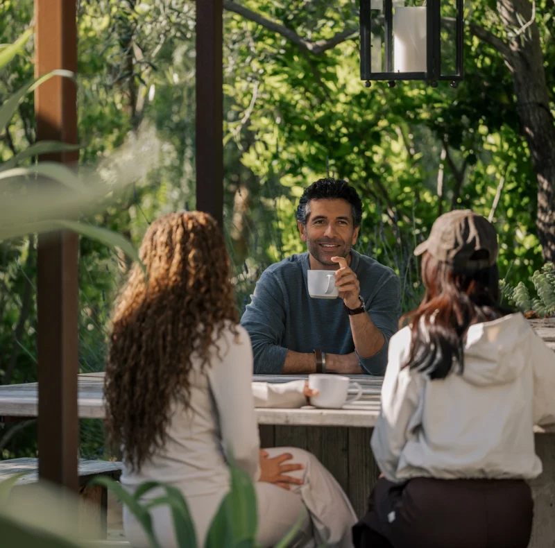 Group of people seated outdoors around a wooden table, chatting and sipping coffee in a sunny garden setting.