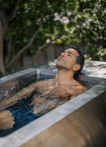 Man with eyes closed rests in an outdoor metal tub filled with water, surrounded by greenery in the background.