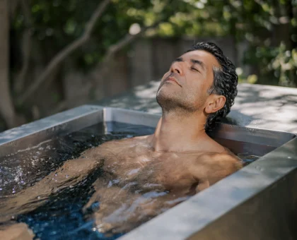 Man with eyes closed rests in an outdoor metal tub filled with water, surrounded by greenery in the background.