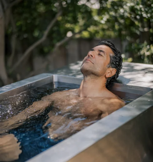 Man with eyes closed rests in an outdoor metal tub filled with water, surrounded by greenery in the background.