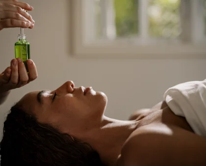 Person lying down receiving a facial treatment; a practitioner applies serum with a dropper from a green bottle