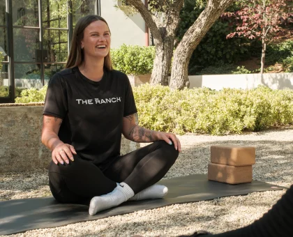Smiling woman in a black T‑shirt sits cross‑legged on a yoga mat outdoors, with trees and a building behind her.