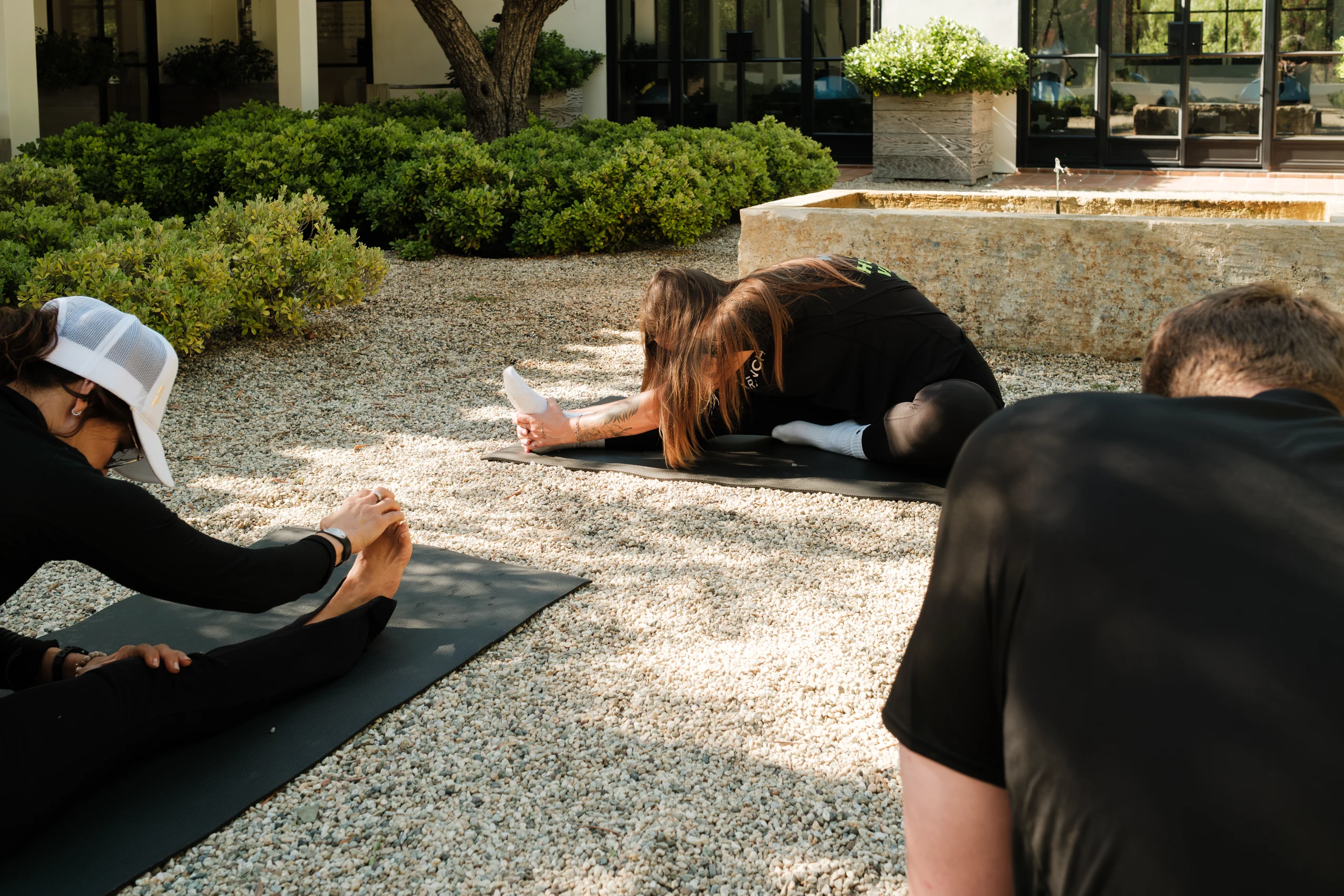 Group of people in black clothing practicing yoga on black mats outdoors, bending forward to touch their toes amid gravel and shrubs near a modern building.